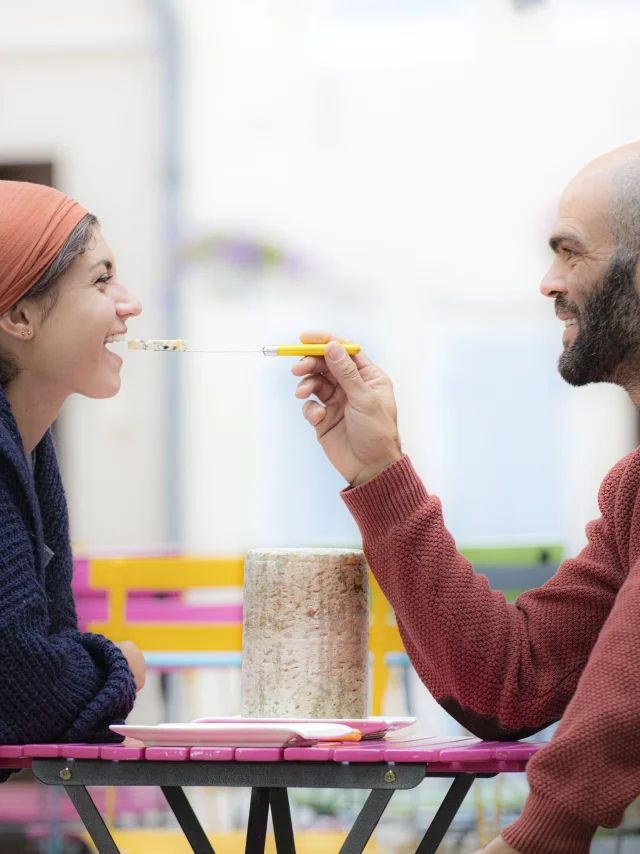 Couple dégustant de la Fourme d'Ambert à une table de jardin, l'homme offre un morceau de fromage à la femme.