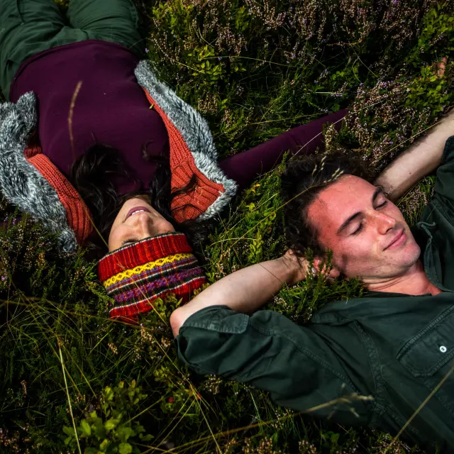 Homme et femme allongés dans les hautes chaumes, entourés de bruyères, profitant d’un moment de détente.