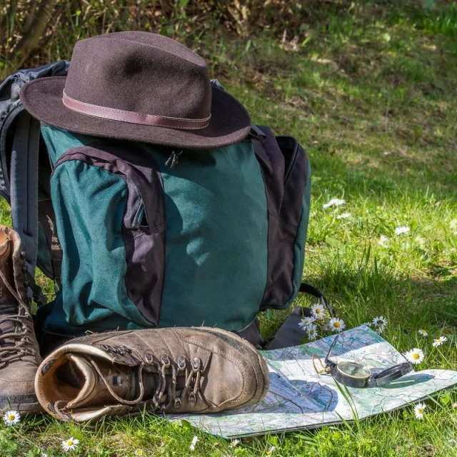 Équipement de randonnée comprenant un sac à dos, des chaussures, une carte et une boussole, posé sur une pelouse fleurie.