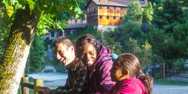 Une famille souriante contre une barrière en bois avec, en arrière-plan, le Moulin Richard de Bas bien éclairé, entouré de verdure et longé par un chemin.