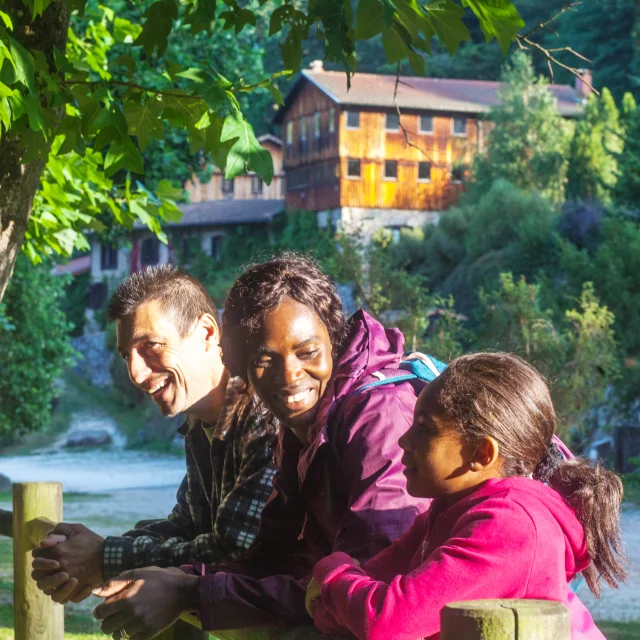 Une famille souriante contre une barrière en bois avec, en arrière-plan, le Moulin Richard de Bas bien éclairé, entouré de verdure et longé par un chemin.