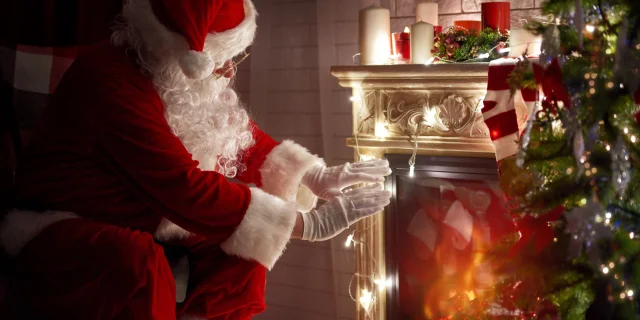 Un Père Noël traditionnel se tient près d'une cheminée décorée de bougies et de lumières de Noël, en train de se réchauffer les mains dans une atmosphère cosy.