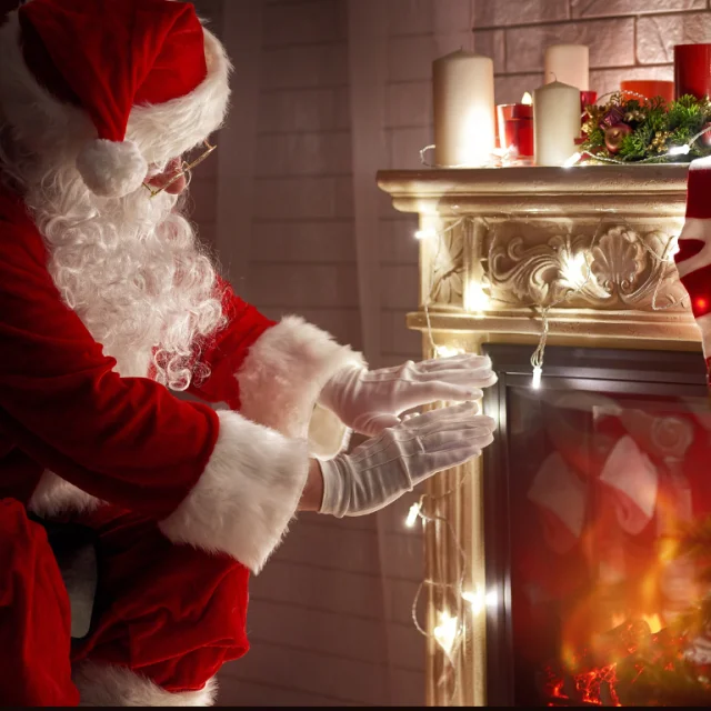 Un Père Noël traditionnel se tient près d'une cheminée décorée de bougies et de lumières de Noël, en train de se réchauffer les mains dans une atmosphère cosy.