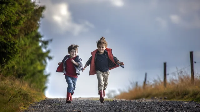 Deux enfants, un garçon et une fille, courent sur une route entourée de sapins et de clôtures en bois.