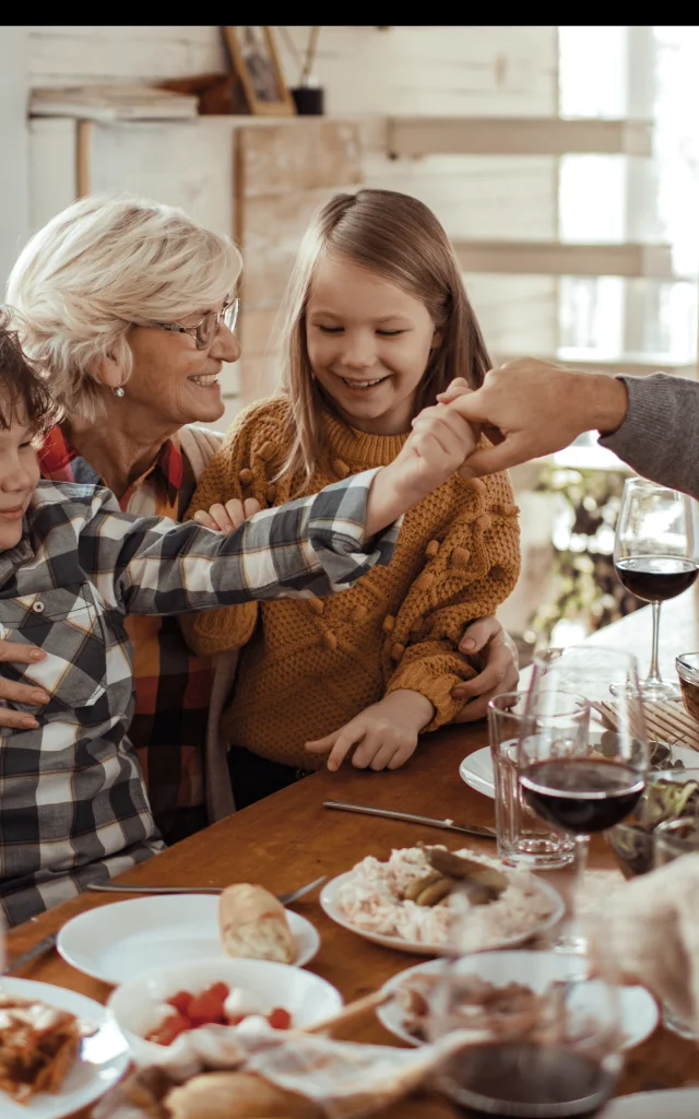 Illustration d’un repas de Noël en famille avec une grand-mère souriante entourée d’enfants, un grand-père et des parents autour d’une table festive en bois.