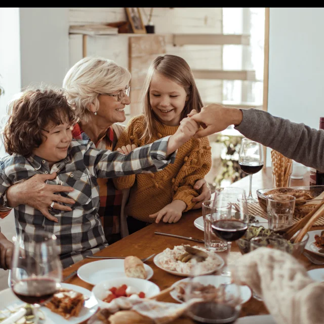 Illustration d’un repas de Noël en famille avec une grand-mère souriante entourée d’enfants, un grand-père et des parents autour d’une table festive en bois.