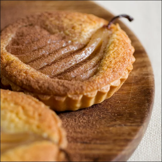 Gros plan sur une tartelette de poire décorative, avec des tranches de poire et une queue de fruit visible, posée sur un support en bois.