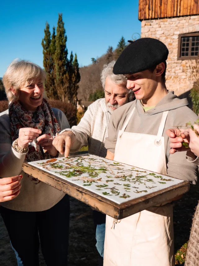 Présentation des feuilles au Moulin Richard de Bas, avec un artisan en béret et tablier, et un couple intéressé par le processus de fabrication du papier.