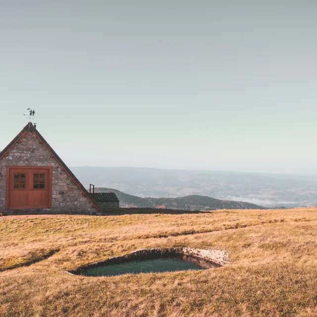 Paysage du Col des Supeyres avec une petite bâtisse en pierre, une mare d'altitude et une vue dégagée sur les montagnes et vallées environnantes.